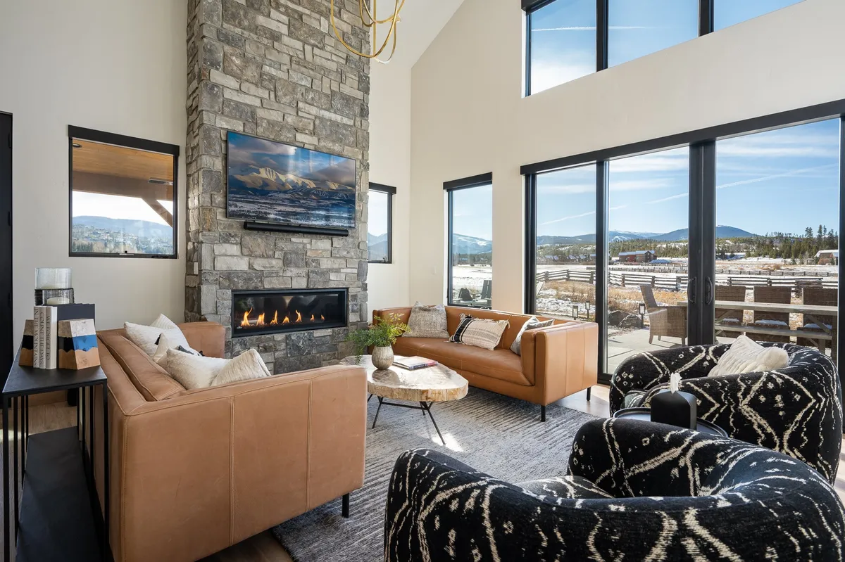 A modern yet cozy living room featuring a stone fireplace, large windows, ample seating, a decorative table, and a large TV mounted above the fireplace. The space is designed for socializing, relaxing, and enjoying both fireside warmth and entertainment.