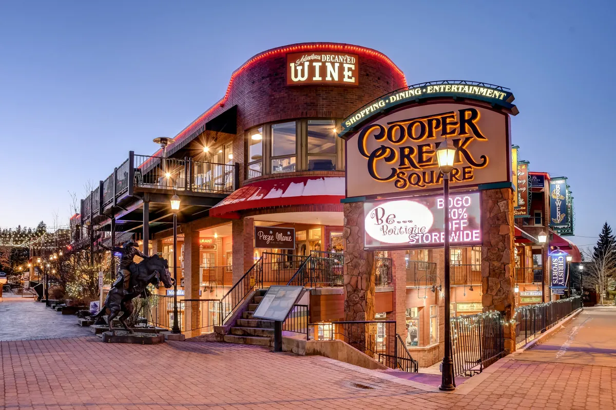 This vibrant exterior scene captures the Cooper Creek Square sign, the entrance to a wine shop, outdoor seating areas, and charming decorative lighting. The bustling atmosphere suggests a lively community hub with shopping, dining, and socializing options right outside your door.
