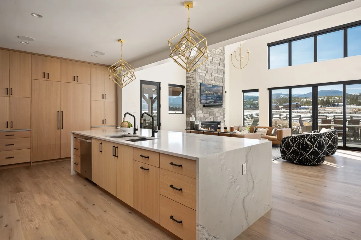A spacious, open-concept living area featuring a large kitchen island with hanging pendant lights, a cozy stone fireplace, expansive windows showcasing mountain views, and a comfortable seating area. The kitchen seamlessly connects to the living and dining spaces, emphasizing togetherness and warmth.