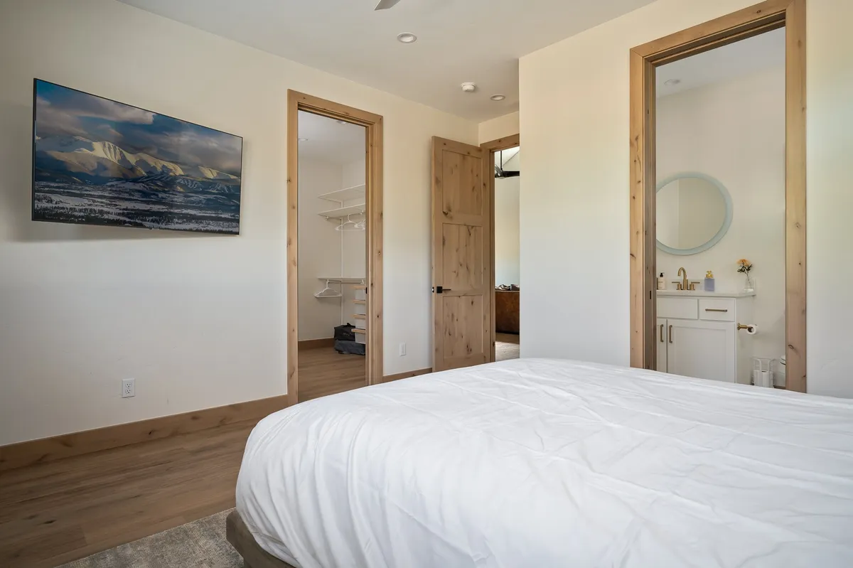 A tranquil bedroom with a bed dressed in crisp white linens, a wall-mounted TV for entertainment, and open doorways leading to an en suite bathroom and closet. The space exudes comfort and privacy, reflecting a restful retreat.