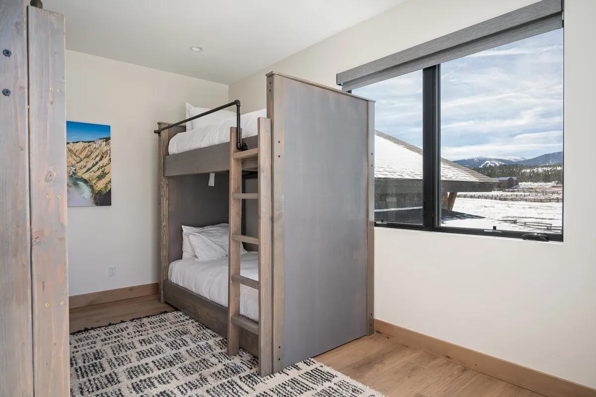 A playful bunk room with two twin-over-twin bunk beds, a large window letting in natural light, and cheerful artwork on the wall. The room is designed to accommodate groups or families, with space for sleeping, reading, and playing games.