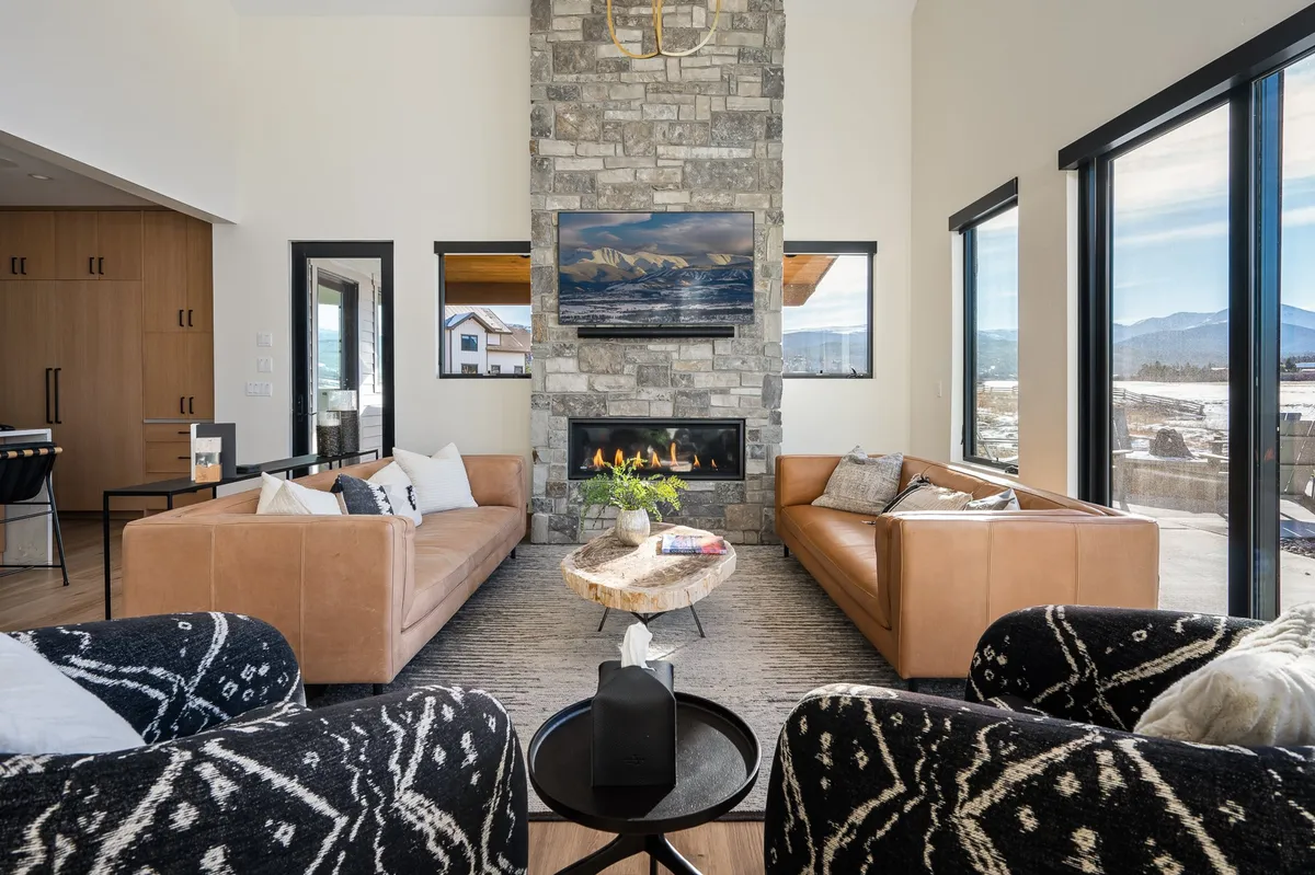 This inviting interior features a cozy stone fireplace, large windows that let in plenty of natural light, plush leather sofas, and warm decor. The space is clearly designed for gathering, playing games, or watching movies together.