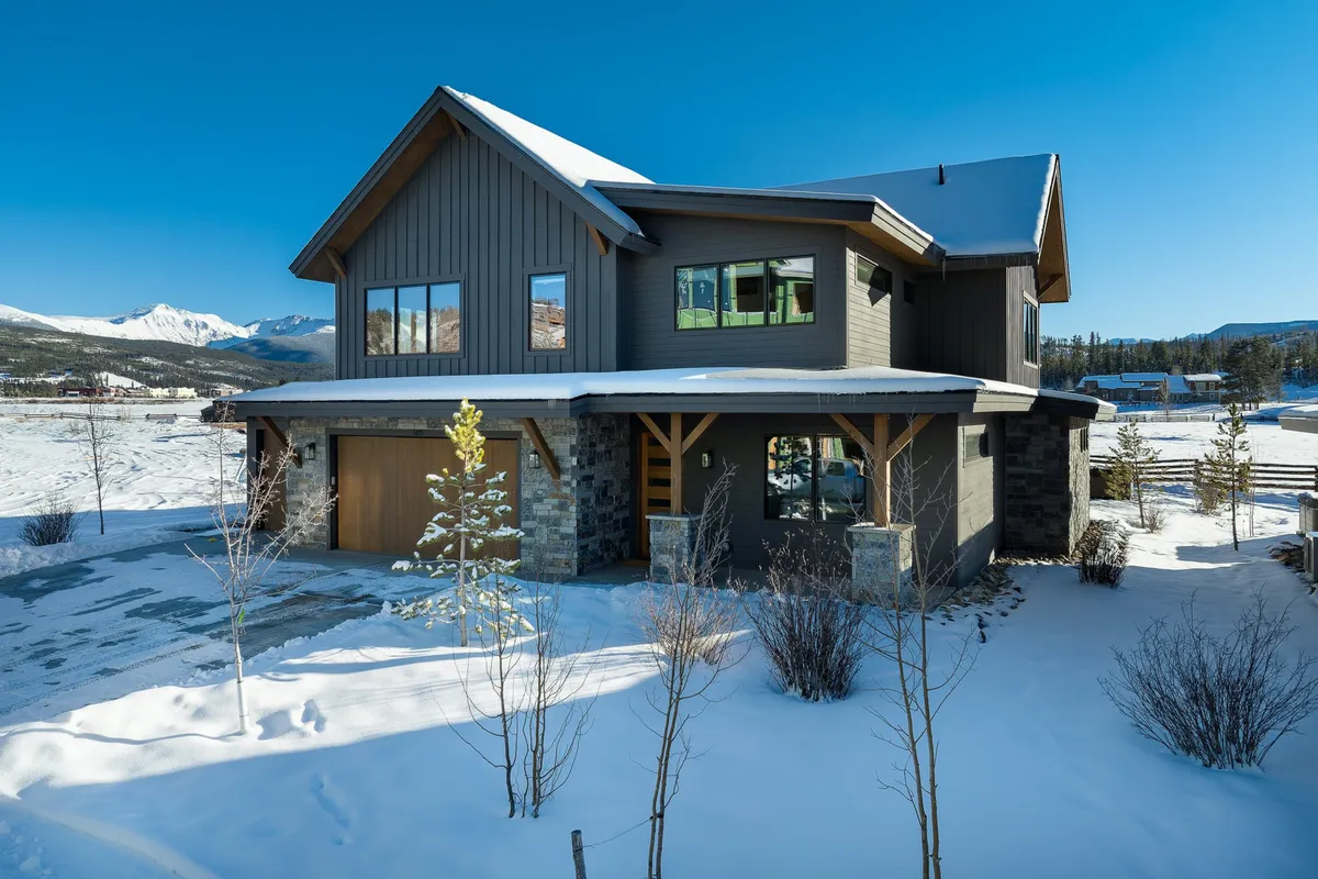 The image showcases the welcoming front entrance of a spacious luxury home, featuring large windows that reveal glimpses of a warm interior. The house sits amid a snow-covered landscape, evoking a sense of winter adventure and cozy comfort.
