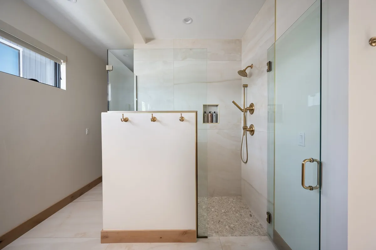 A spa-inspired bathroom with a glass shower enclosure, elegant gold fixtures, minimalist design, and a large window. The space feels luxurious and tranquil, offering a private oasis for relaxation and self-care routines.