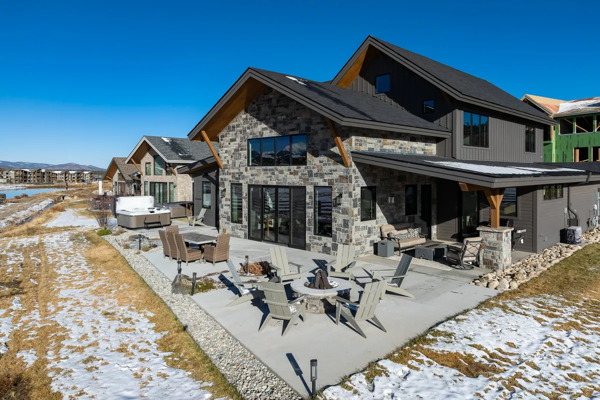 The image showcases the home's exterior with a stone facade, large windows, an inviting outdoor seating area, a fire pit, and a jacuzzi. The patio area is designed for socializing and relaxing, while the natural backdrop connects guests to the mountain scenery.
