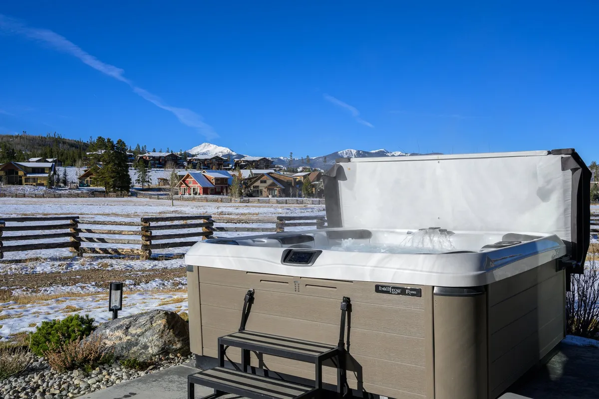 A steaming hot tub sits on a wooden patio with snow-covered ground and mountains in the background, inviting guests to relax while taking in the winter scenery. A wooden fence frames the space, creating a sense of privacy while still offering panoramic views of nature.