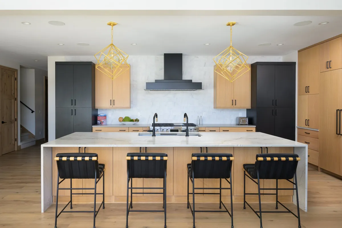 A modern kitchen scene with a prominent island countertop, elegant gold pendant lights, sleek black cabinetry, and high-end stainless steel appliances. The setting exudes sophistication and is ideal for culinary creativity and entertaining guests.
