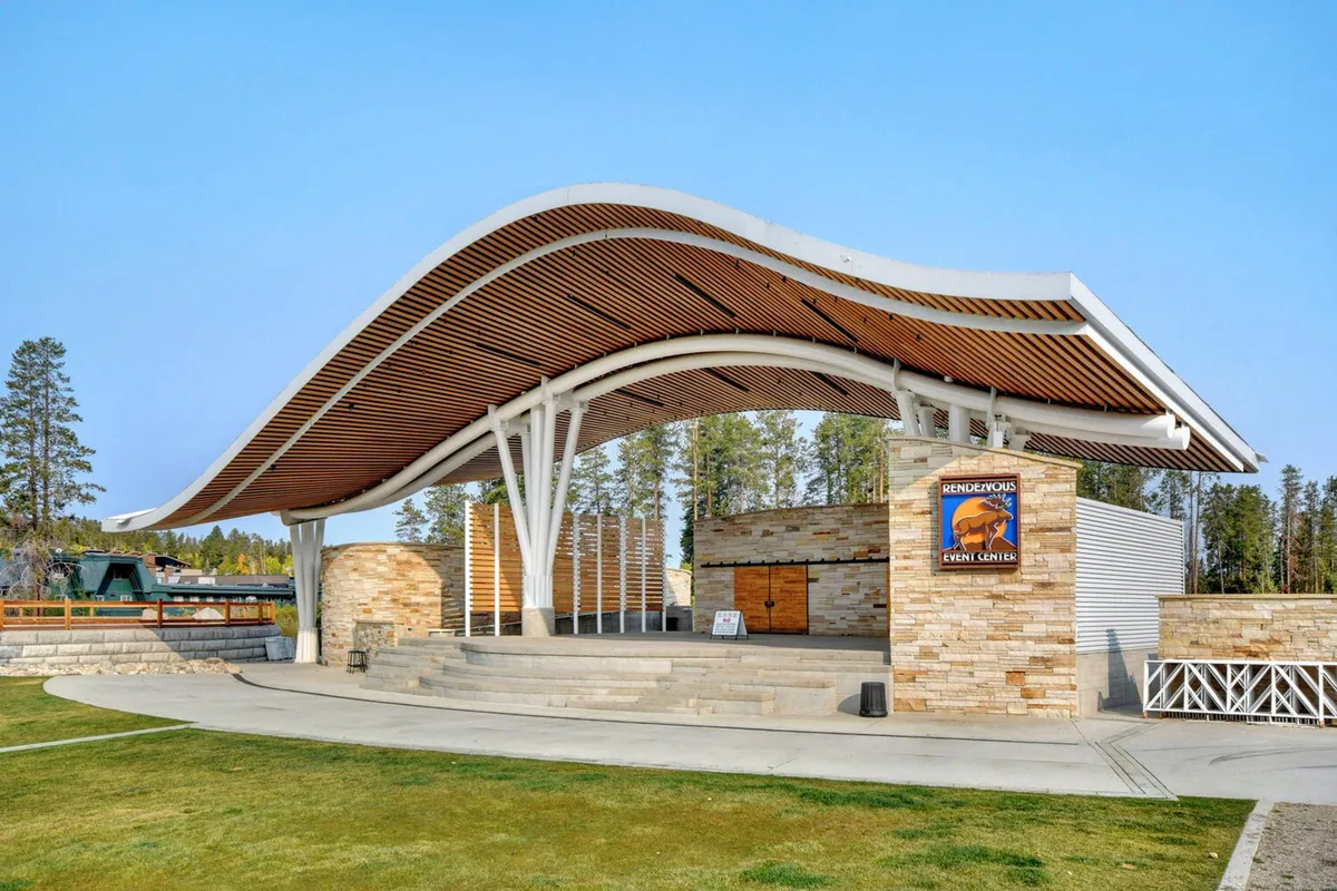 A striking outdoor amphitheater with a curved roof structure and stone walls, featuring steps that lead to a stage. The setting is built for concerts, festivals, and community gatherings, conveying a sense of lively outdoor events and local celebration.
