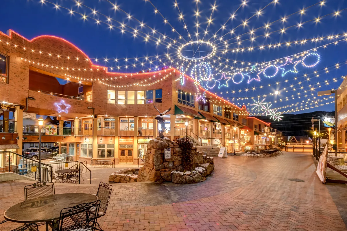 A lively evening scene of a mountain town with decorative lights, a stone fountain, outdoor seating, and buildings with large windows. People are dining, shopping, and socializing in a festive atmosphere, reflecting the area's year-round vibrancy.